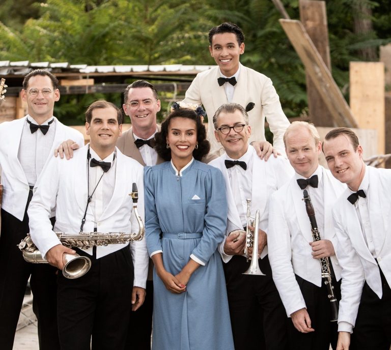 Gruppe von sieben Musikern in Anzügen und einer Frau in einem blauen Kleid, lächelnd.